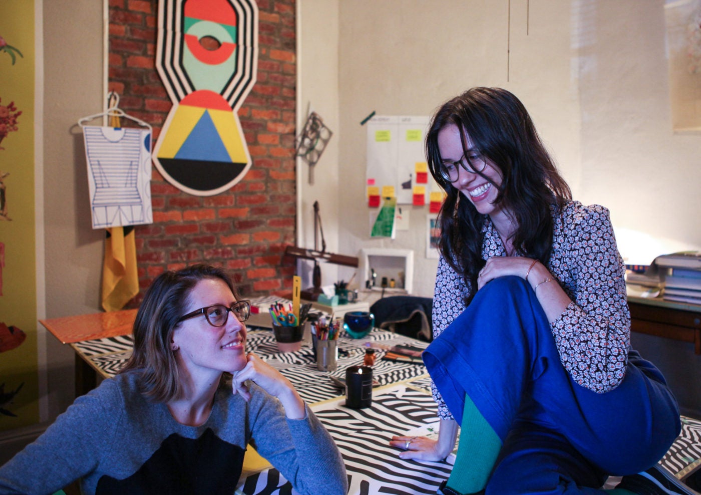 two white women with brunette hair and glasses sit together looking at eachother