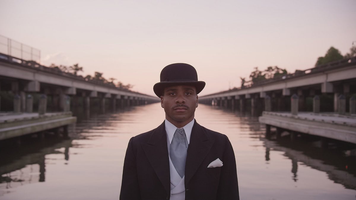 Black man in a bowler hat and suit, with a water canal receding in the background, from a John Akomfrah film called Precarity.