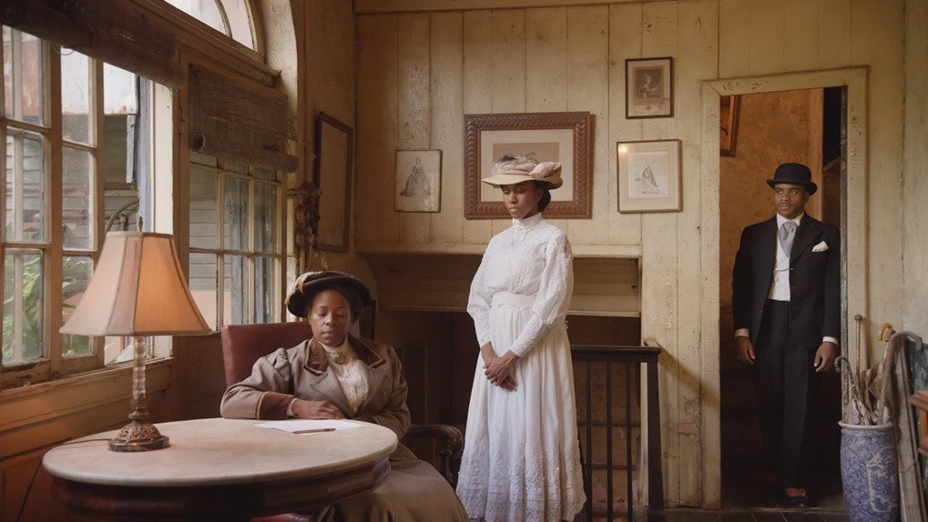 A black woman sits at a table, and a black woman stands next to her, and a black man in a bowler hat stands in the doorway, all in 19th-century attire and setting. From a John Akomfrah film.