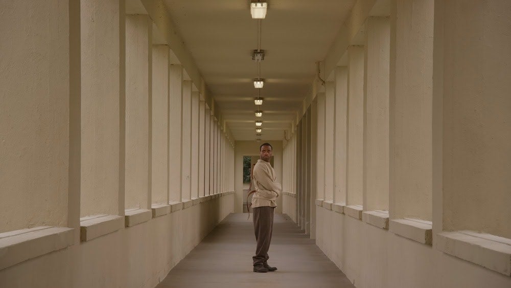 Black man in a strait jacket standing in a corridor looking at viewer. From John Akomfrah film Precarity.