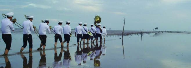 A line of 10 musicians marching in a flooded street with a tuba leading the way, seen from behind. 