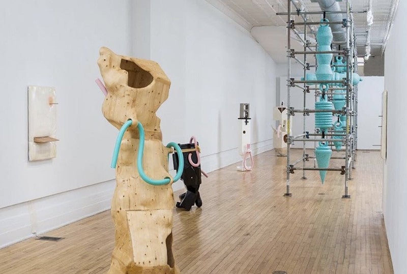 A long narrow room with a wood floor and various styles of sculptures installed on the floor with shared sea foam green and pink color accents.