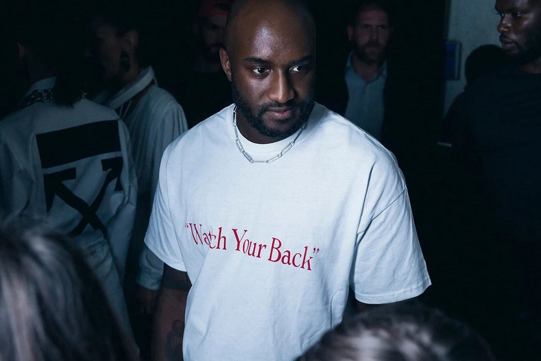 image of a dark skinned man with a goatee and bird wearing a short paper clip chain and a white t-shirt with red letters that say "Watch Your Back," he is spotlit in a crowded dark room