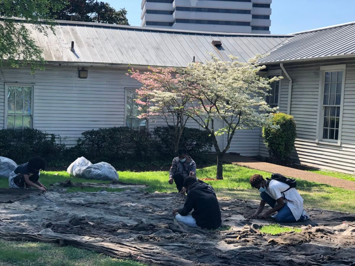 four people sit on the grass sewing burlap sacks. 
