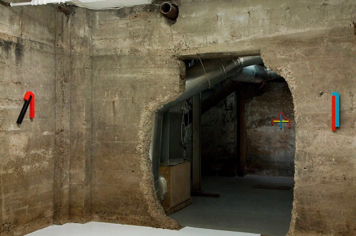 Small multicolor shaped sculptures hang in a cement gallery with strange architecture