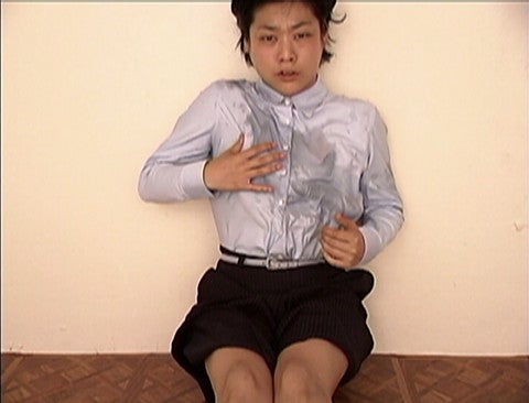 still from a film, an Asian woman with a distressed facial expression sits on the ground with their backing against a white wall, wearing a white button down and black pinstripe shorts, the chest of the shirt is wet with what looks like water