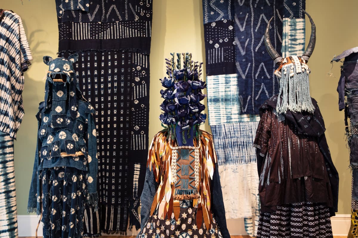 five elaborately masked figures/icons in dark blue, gold, and neutral colored costumes stand in a v-formatio in a room with elaborate navy and white curtains and light yellow-green walls