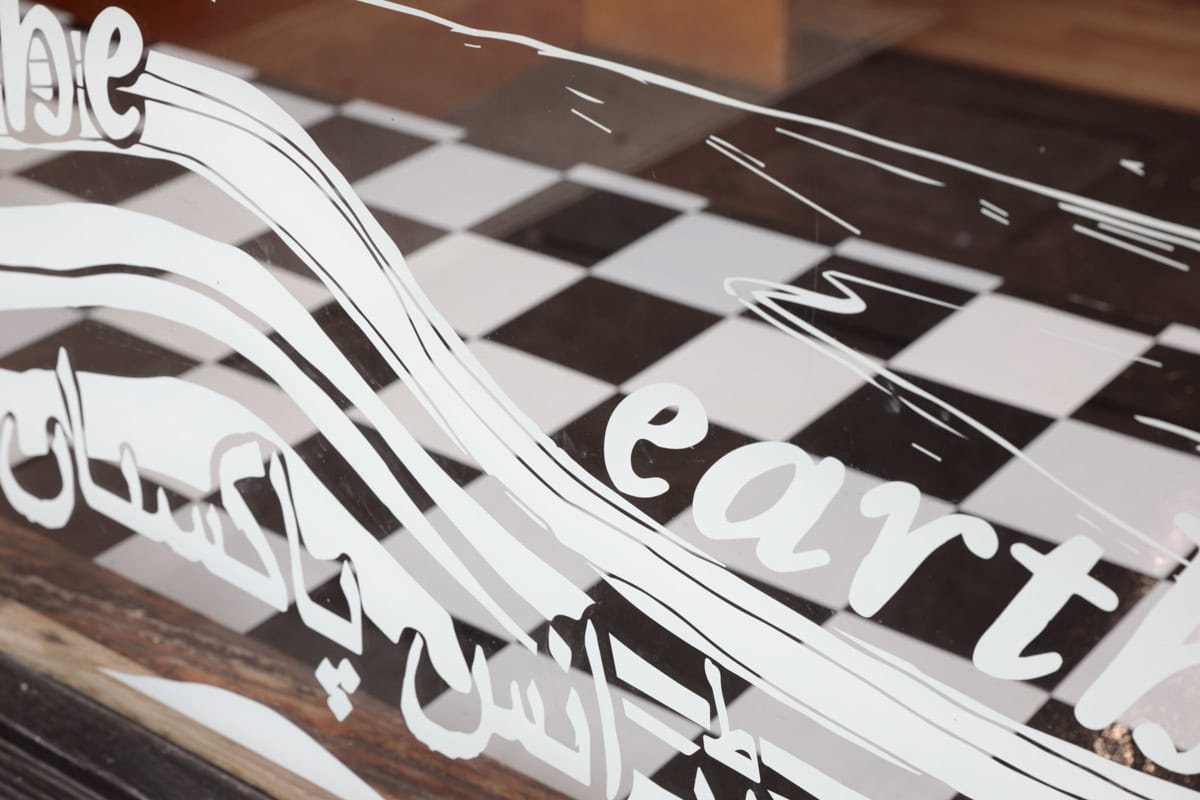 a mirror with the exhibition title written in white cursive English text and Urdu script. the mirror reflects a black and white checkered title floor and dark brown wooden accents in the exhibition space