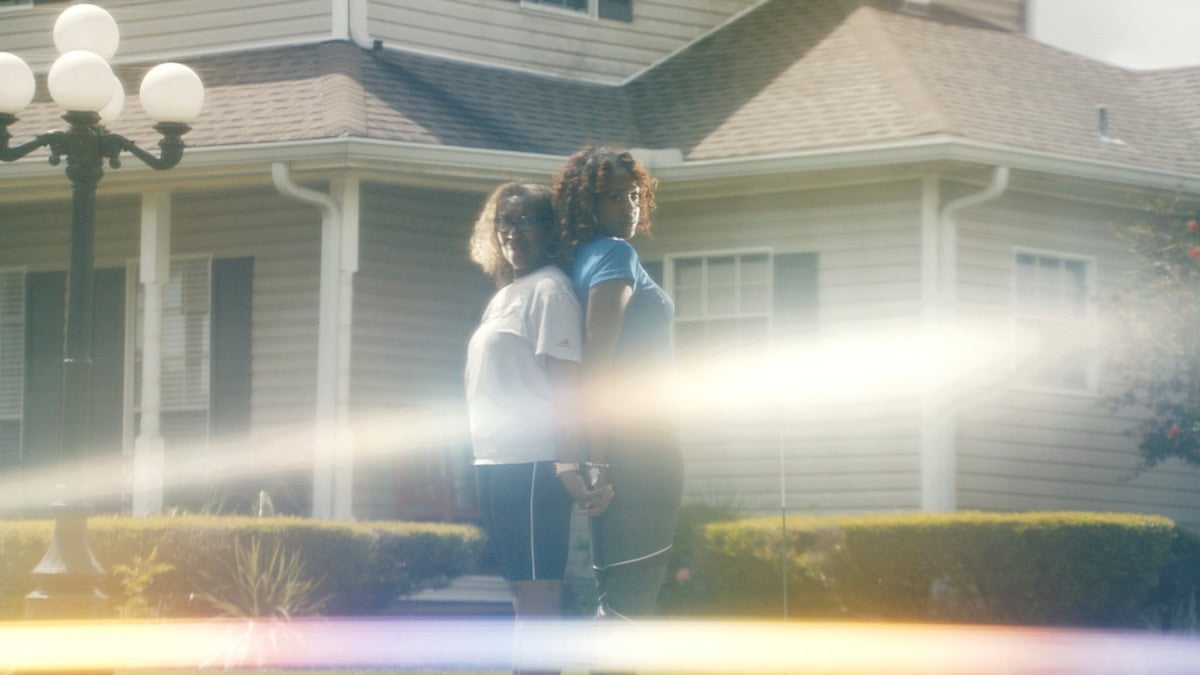 a older Black woman and younger Black woman stand back-to-back in front of a white house, they look proud and determined, staring into the camera as a rainbow light underlies the photograph