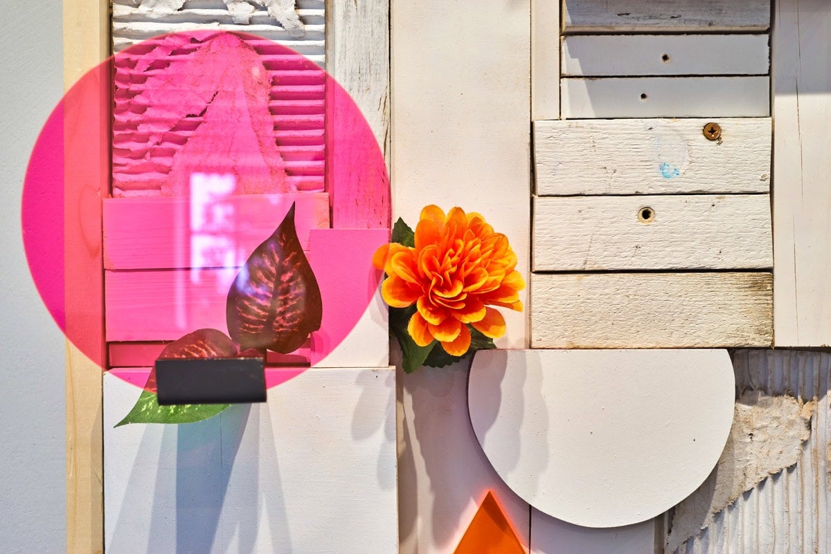 A detail view of a vertically horizontal altarpiece-like structure made of light blonde wood pieces and white geometric shapes. There is a transparent neon pink circle with two green teardrop-shaped leaves seen behind it. There is a bright orange flower with green leaves at the center of the image.