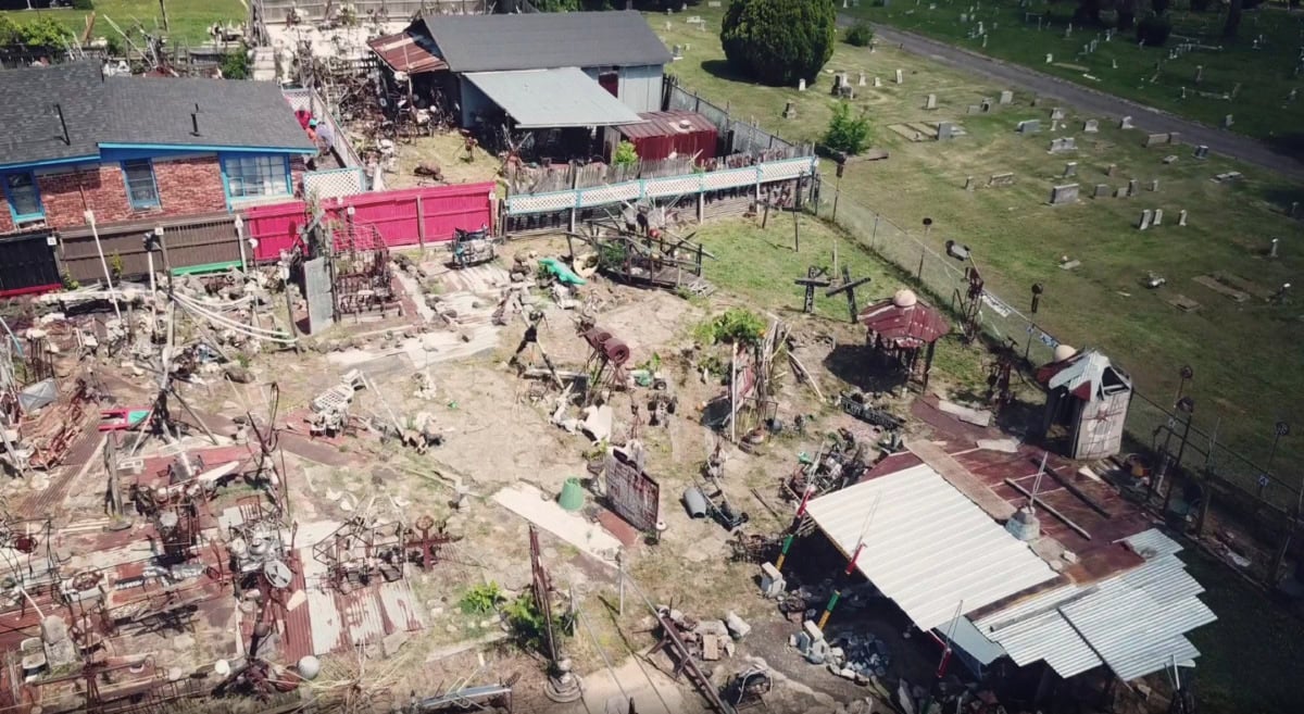 an aerial shot of a multi-acre yard show with collections of metal, painted signs, and sculptural artifacts in careful, yet overwhelming proportions, on the right side of the yard is a cemetery of green grass and humble gravestones