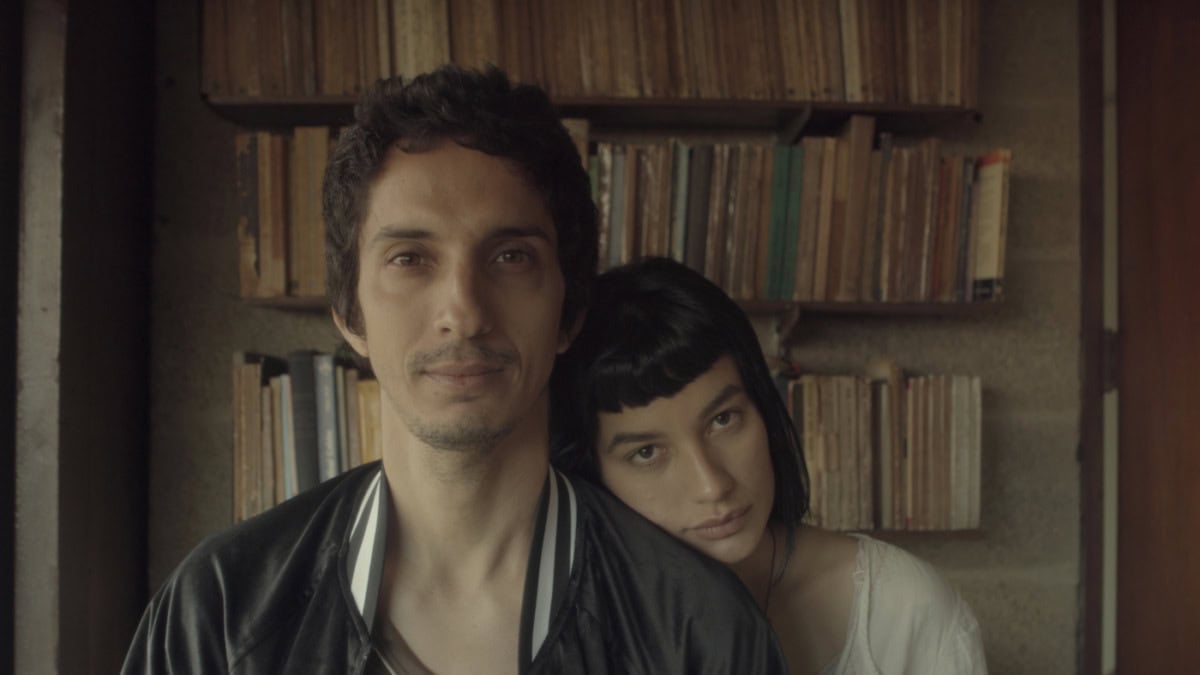 a white couple looks directly at the camera. a large bookcase looms behind them, the woman has her head on the man's shoulder. they have soft smiles on their face