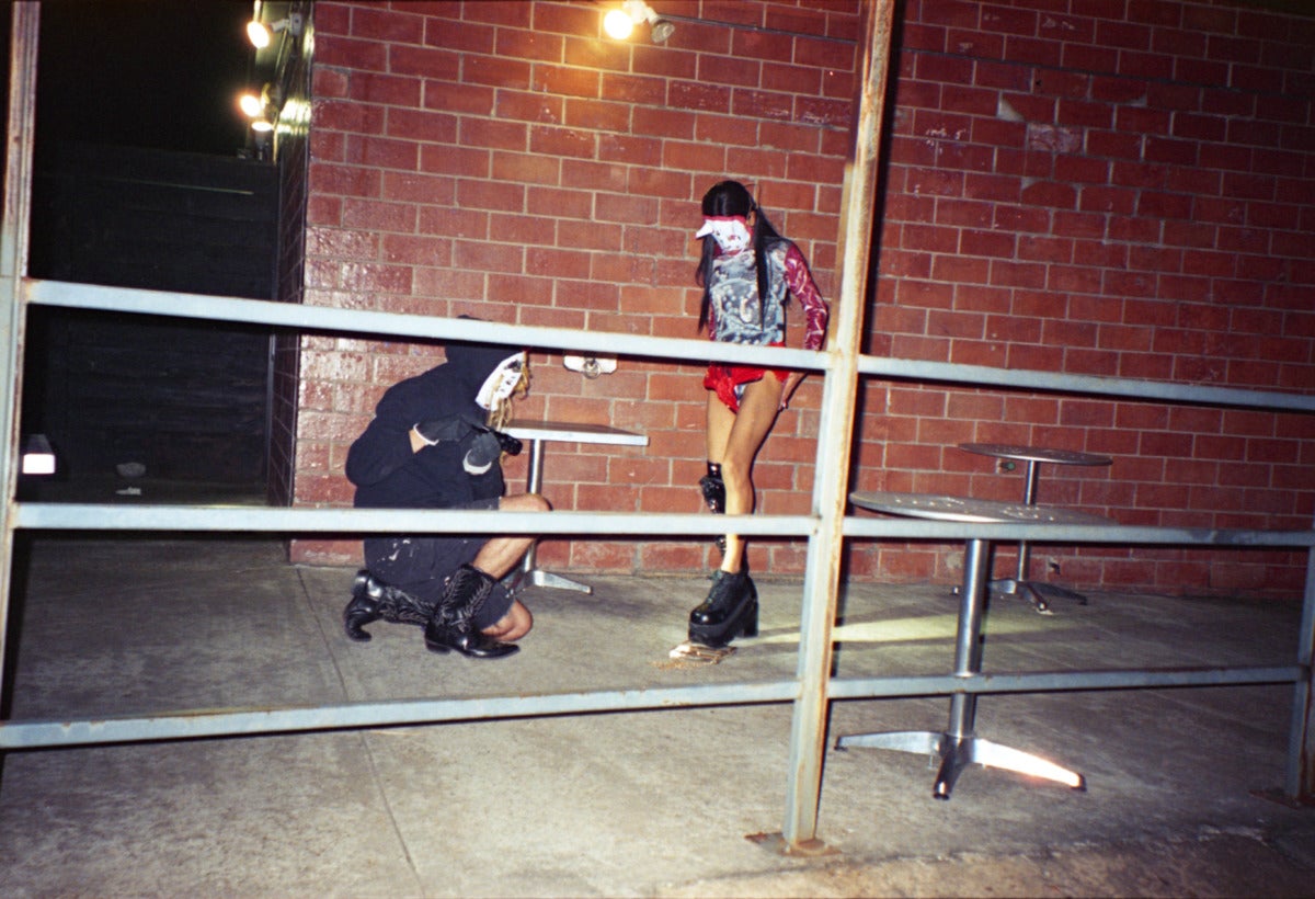 two Kabuki masked people stand in front of a brick wall at night. the person on the left is wearing all black and cowboy boots, shining a flashlight at the platform black boots of the person on the right. the person on the right has long hair and is wearing red shorts. the person on the right is crushing a gold purse under their foot. 