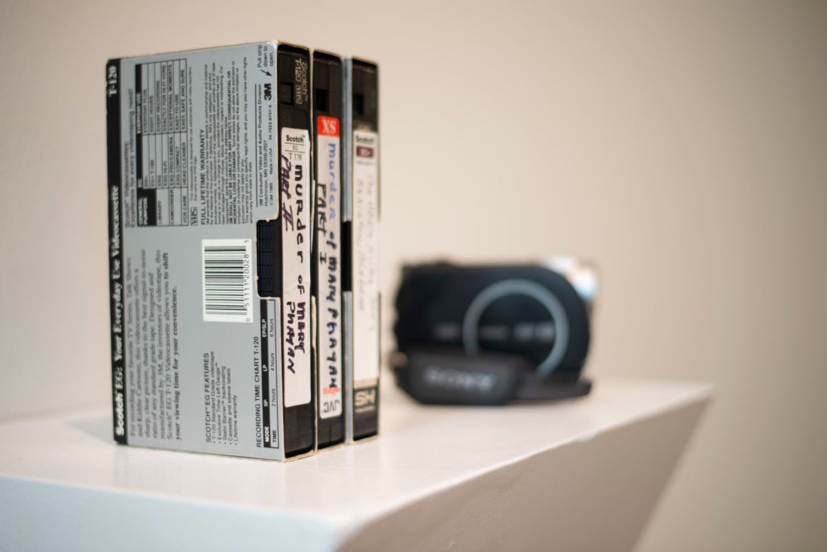 on a white podium against a white wall are three stacked VHS tapes of home made movies. the labels on the VHS tapes say the murder of Mary phatah parts 1-3. In the background there is a handheld camcorder.