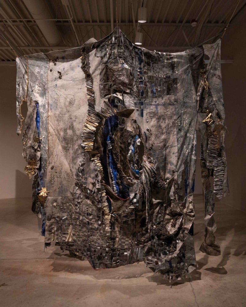 a large grey and beige colored tapestry with blue ripples and ruffles hangs in the middle of the gallery.