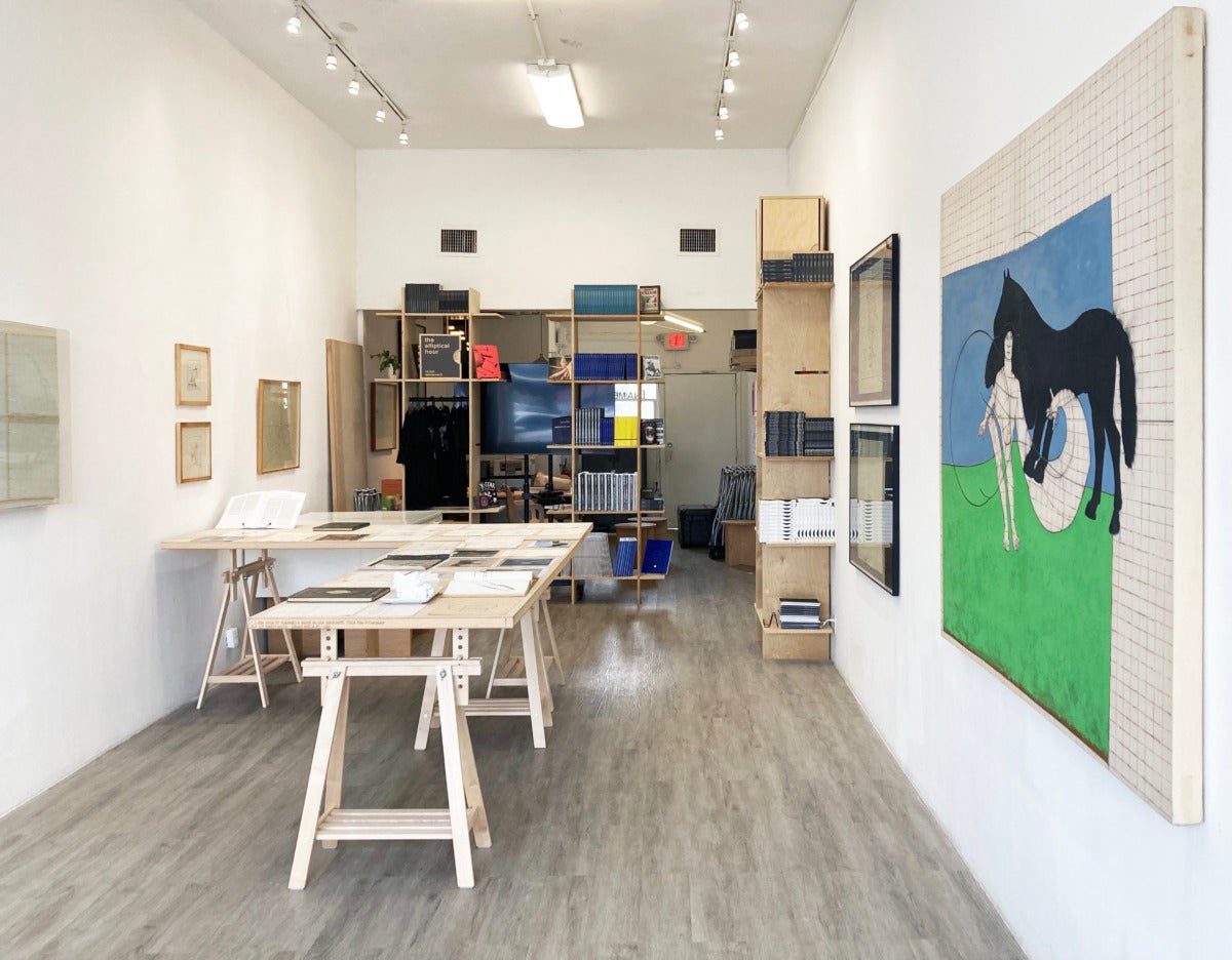 a small white walled space with a light wooden L shaped table on saw horses covered in drawings on paper. on the wall there is a large grid painting of a black horse and a white figure. at the back of the room are light wooden shelves with books.