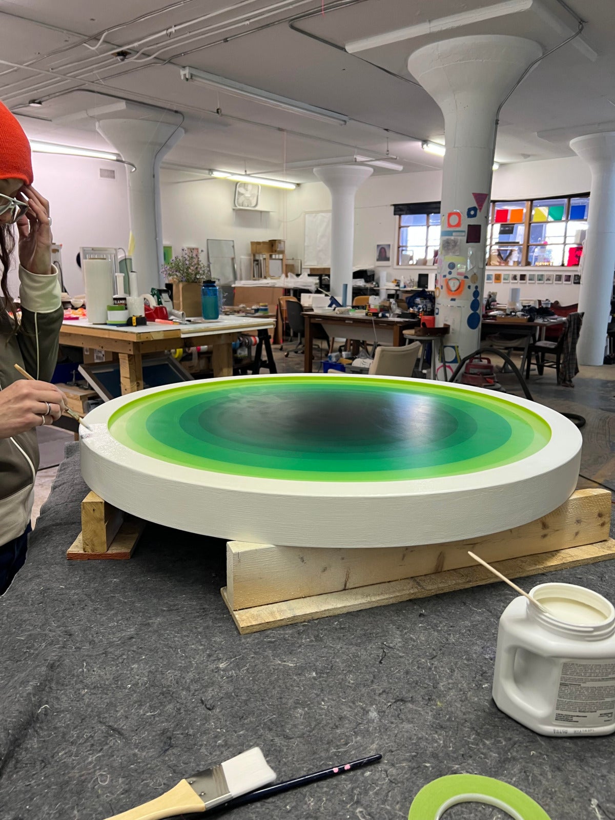a busy studio with large white pillars, with tables covered in art supplies. in the foreground, a white woman with an orange beanie and rings on her hands works on a large circular painting with reverberating shades of green