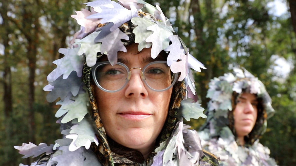 A figure wearing glasses faces the camera. An additional figure is obscured in the background. Both are wearing hoods covered in artifical flowers.
