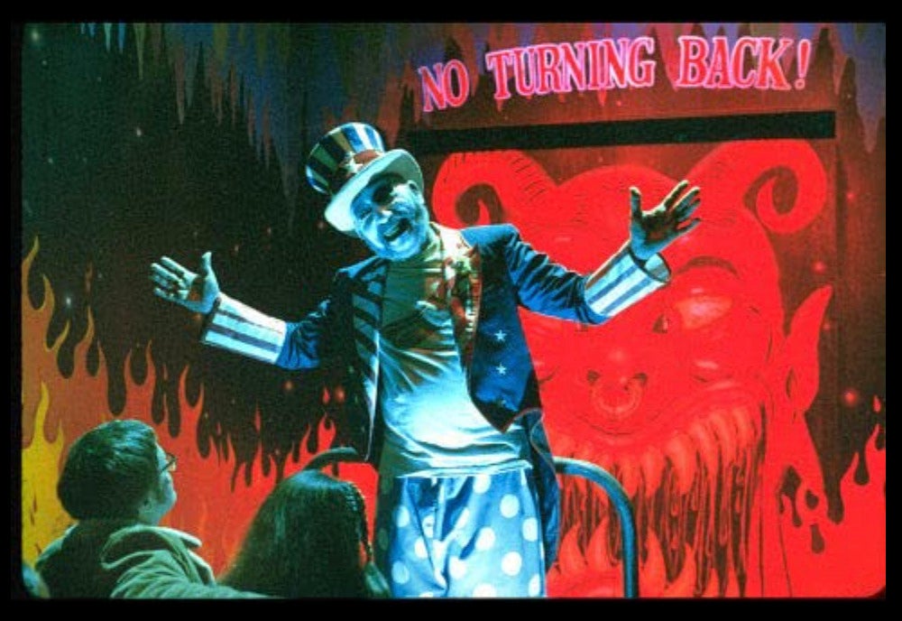 A man dressed in an Uncle Sam costume with a menacing smile greeting two people at an amusement park ride