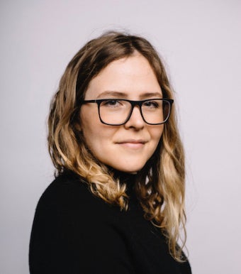 a headshot of Laurel V. McLaughlin, who wears a black turtleneck against a light grey backdrop. Laurel has brown sun-bleached hair and wears large square glasses