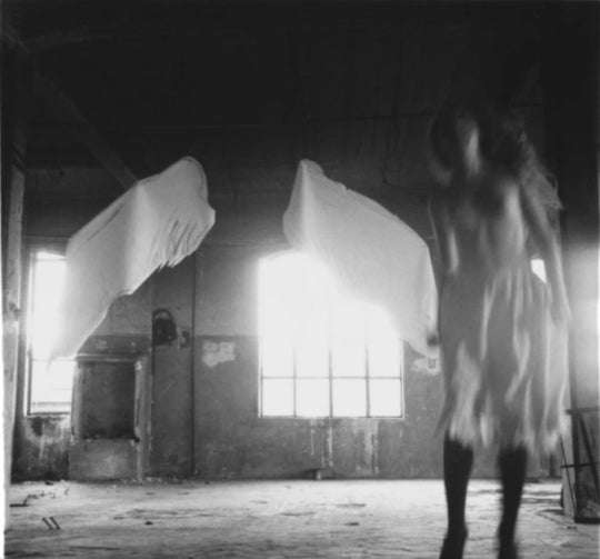 an ethereal black and white portrait of a woman wearing a white dress in a large sunlit room, with shadows bouncing across the space