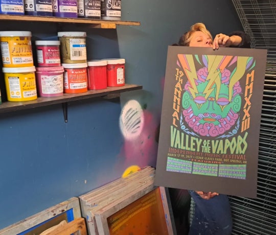 standing in front of a shelf lined up with paint, woman holds poster up of a music festival that took place in Arkansas