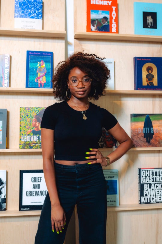 a Black woman wears a cropped black t-shirt and dark pants with a necklace. her bright manicured hand rests on her hip. she stares at the camera with a thoughtful gaze with wide-rimmed glasses and a curly brown fro
